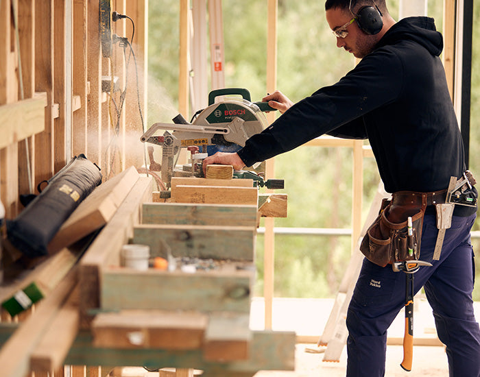 Man sawing wood indoors wearing safety goggles, a toolbelt and Hard Yakka workwear