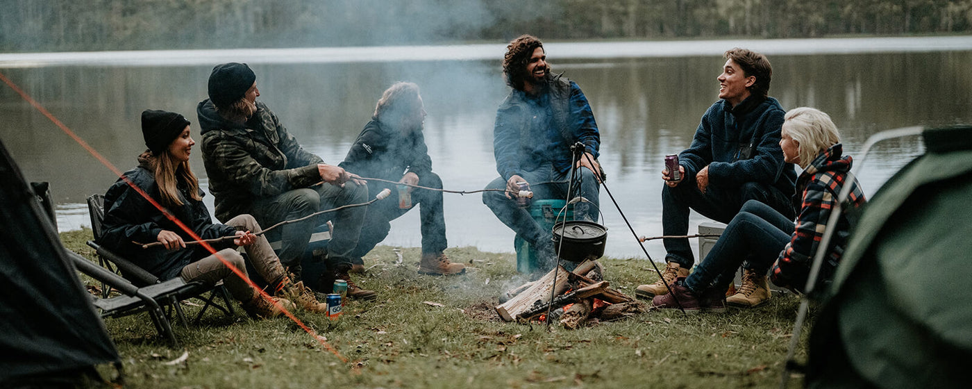 Group of people sitting around a campfire by a lake, wearing Hard Yakka clothing and footwear.