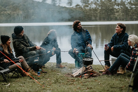 A group of men and women sat around a campfire by a lake, all wearing Hard Yakka workwear.