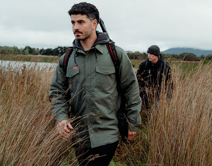 Two men walking through tall grass in a natural setting, the man at the front is wearing a Hard Yakka Solid Shacket.