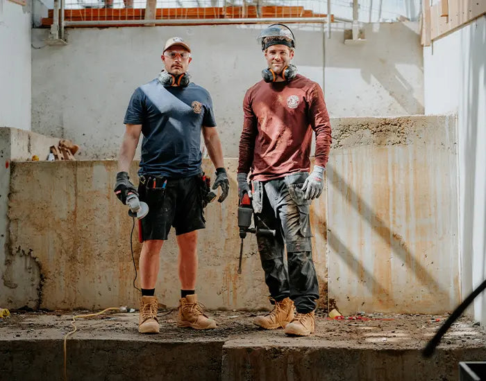 Two men inside a sunlit building holding power tools and wearing Hard Yakka workwear.
